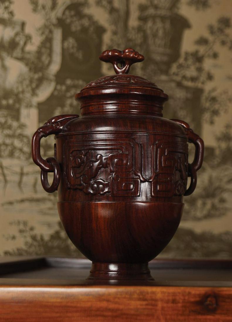 CHINESE HARDWOOD VASE AND COVER: with archaistic carved bank and elephant scroll handles, raised on a circular foot. 20 cm. high; 15 cm. diameter