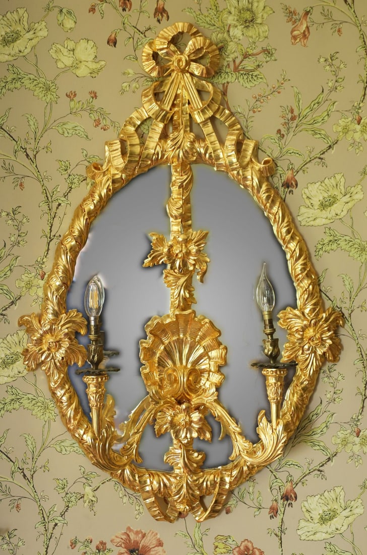SET OF FOUR CARVED GILTWOOD WALL SCONCES: WITH MIRROR PLATES, Each with scrolling foliate arms issuing from a shaped backplate incorporating a mirror plate, the frames richly carved with rocaille and floral ornament, retaining a gilt finish a