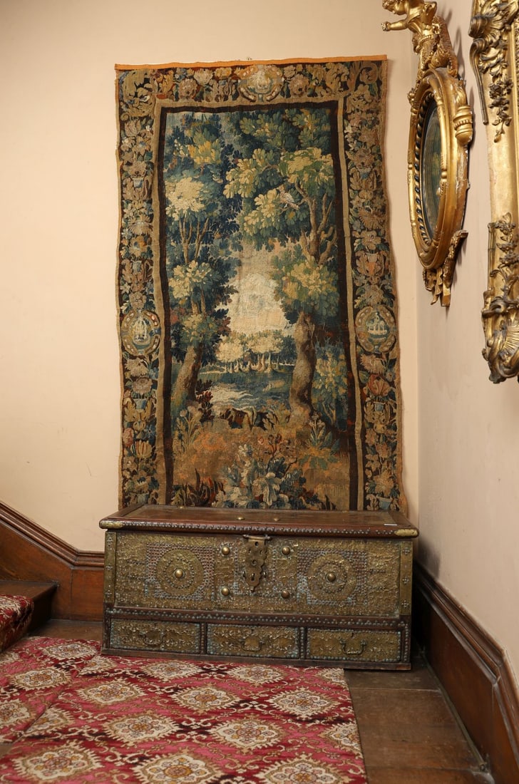 17TH-CENTURY VERDURE TAPESTRY, SILK AND WOOL: A 17th-century verdure tapestry woven in wool with silk highlights, depicting a wooded river landscape framed by flowering trees and dense undergrowth. The vertical composition is structured by two su