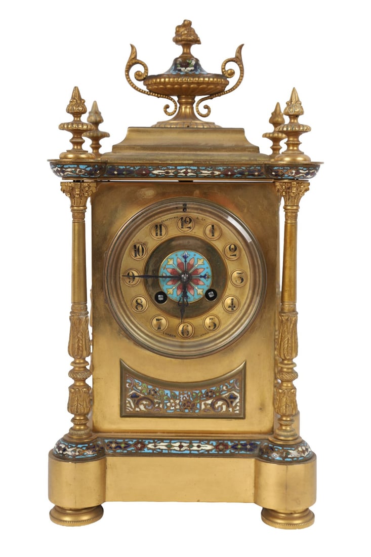 FRENCH GILT-BRONZE &CHAMPLEVÉ ENAMEL MANTEL CLOCK: LATE 19TH-CENTURY. The architectural case surmounted by an urn finial and flanked by column supports, the gilt-bronze body inlaid with polychrome champlevé enamel panels, the circular dial with Arabi