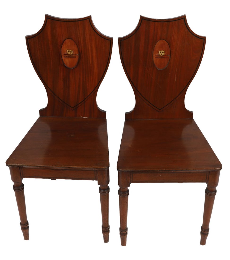 PR GEORGE III HEPPLEWHITE ARMORIAL HALL CHAIRS: A pair of George III Hepplewhite shield-back hall chairs, each with inlaid armorial crest to the back above solid seats and square tapering legs. FURNITURE