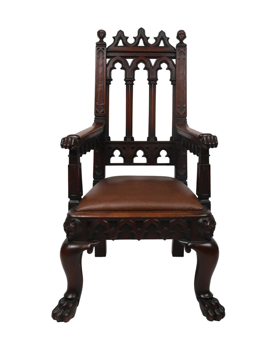 GOTHIC REVIVAL CARVED MAHOGANY ARMCHAIR: A 19th-century Gothic Revival carved mahogany armchair, the high back with pointed arch tracery and pierced panels beneath a crenellated top rail, flanked by uprights surmounted with carved bust finia