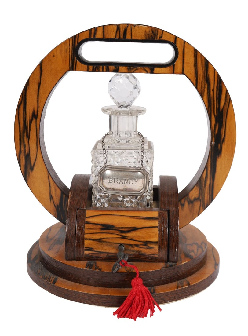 NOVEL 19TH-CENTURY TANTALUS: A late 19th-century novelty tantalus in figured maple and ebonised wood, of circular form with integral handle, enclosing a cut-glass decanter with faceted stopper, the base fitted with a locking mech