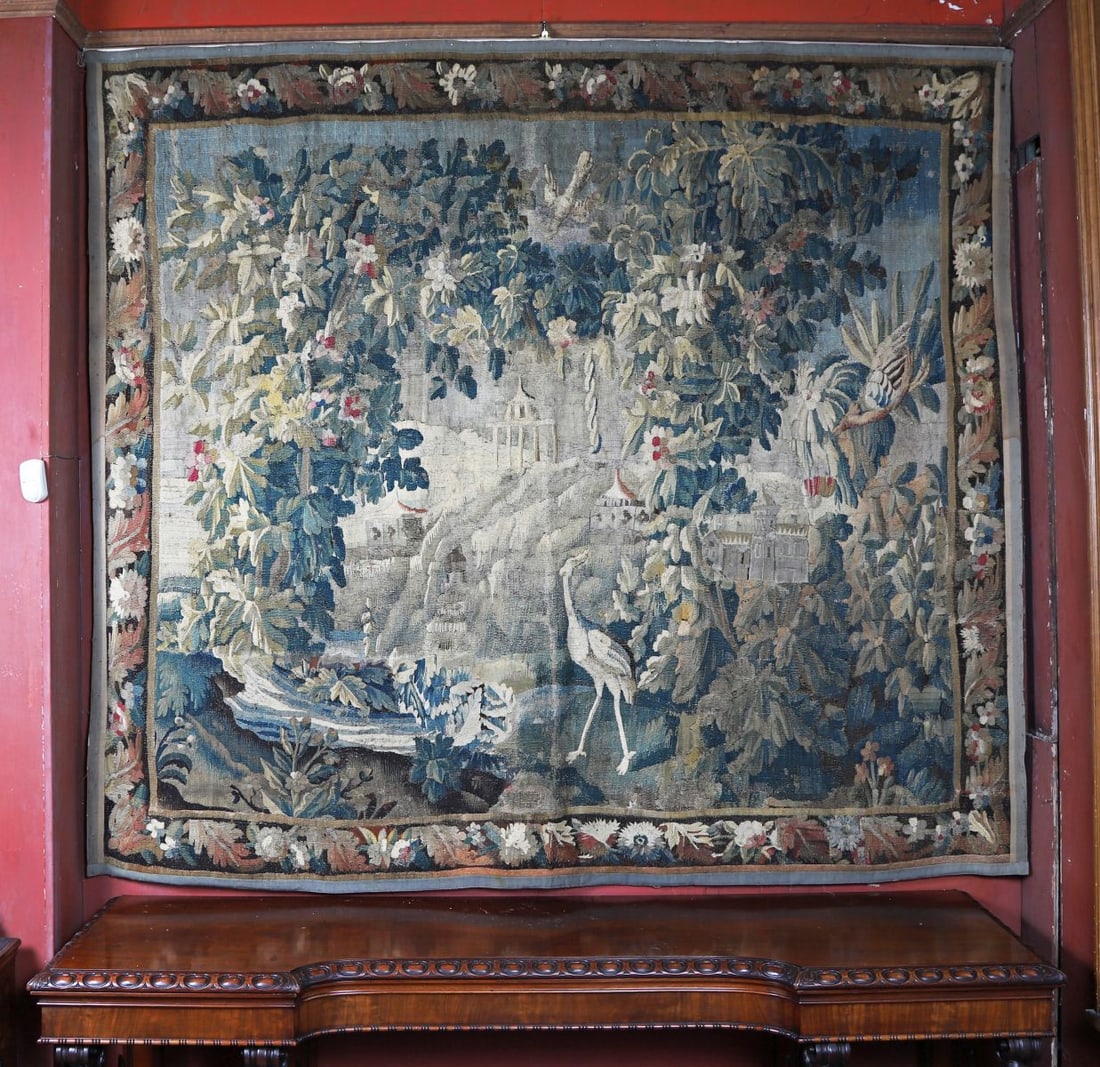 17TH -CENTURY FLEMISH VERDURE TAPESTRY: Large hand-woven silk and wool verdure tapestry depicting a wooded landscape populated with exotic birds, including a crane and parrot, set amidst dense foliage with architectural elements and distant
