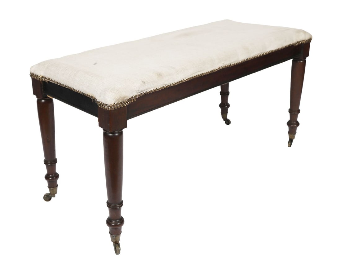 MAHOGANY AND UPHOLSTERED WINDOW SEAT: The rectangular seat with loose upholstered cushions edged with studwork, raised on turned tapering legs with brass caps and castors. FURNITURE