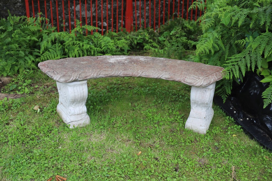 CURVED COMPOSITE STONE BENCH WITH SCROLL DETAIL: A garden bench in composite stone, with a curved top and deeply moulded scrollwork design to the edge, raised on twin scrolled supports. Weathered with red lichen patina.