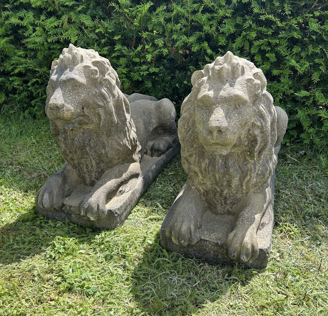 PAIR OF COMPOSITE STONE RECLINING LIONS: Pair of reclining composite stone lions with flowing manes, each modelled recumbent upon a rectangular plinth base. Weathered surface patination.