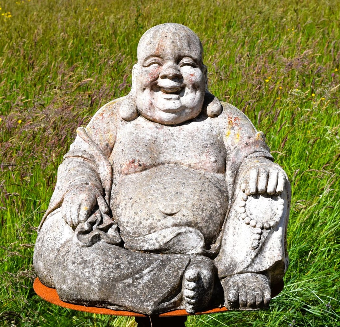 COMPOSITE STONE BUDDHA GARDEN ORNAMENT: Composite stone model of a seated laughing Buddha holding prayer beads and a sack, with weathered patination.