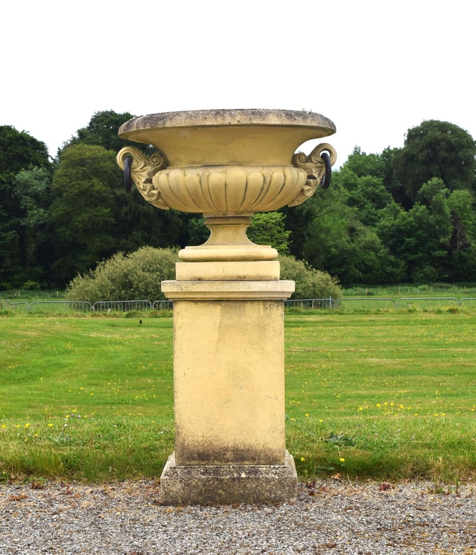 COMPOSITE STONE CAMPANA URN ON PEDESTAL: Composite stone campana urn with ram's mask handles and metal rings, raised on a matching square pedestal.