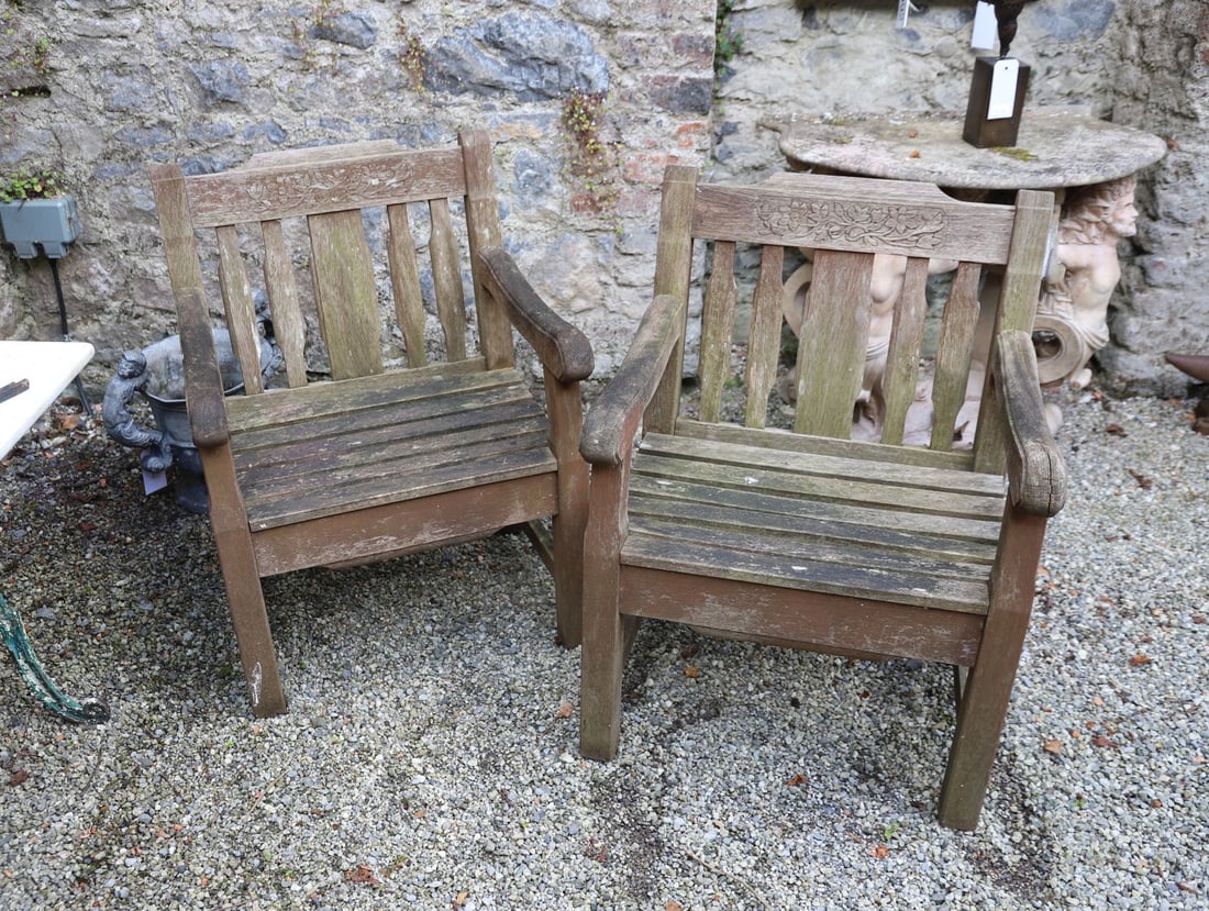 PAIR OF HARDWOOD GARDEN ARMCHAIRS: Pair of weathered hardwood garden armchairs with carved foliate decoration to the cresting rails, slatted seats and shaped arms.