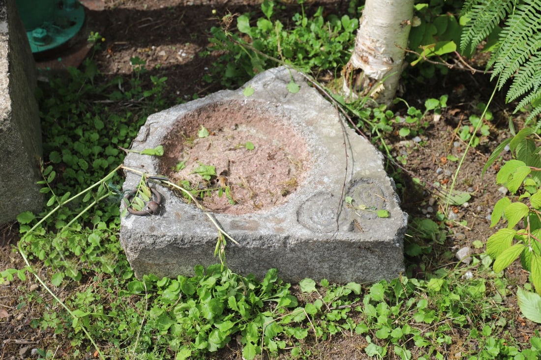 CARVED GRANITE LEAD RING BASIN: A hand-carved granite basin with deep central bowl, featuring an applied iron lead ring and incised decorative spiral motif to the surface. Weathered and mossed
