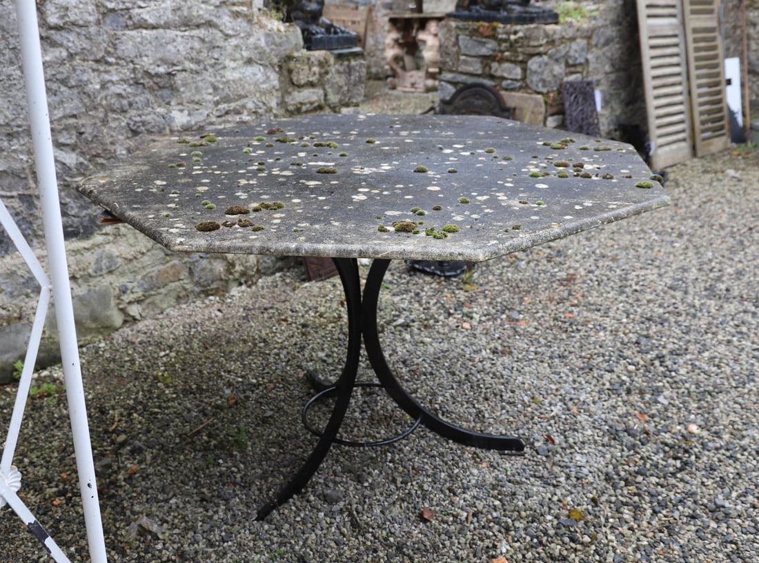 LARGE OCTAGONAL STONE-TOP GARDEN TABLE: Large octagonal stone-top garden table, the substantial weathered stone slab raised on a wrought iron base with splayed legs and circular stretcher. Moss-covered surface with natural weathering.
