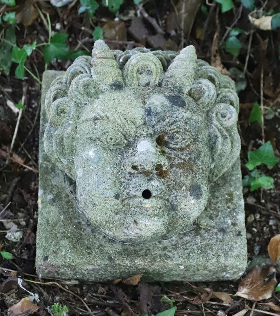 STONE MASK FOUNTAIN HEAD: A carved stone fountain head in the form of a putto, with flowing curls and stylised facial features. Central spout aperture above the brow. Weathered surface with lichen traces.