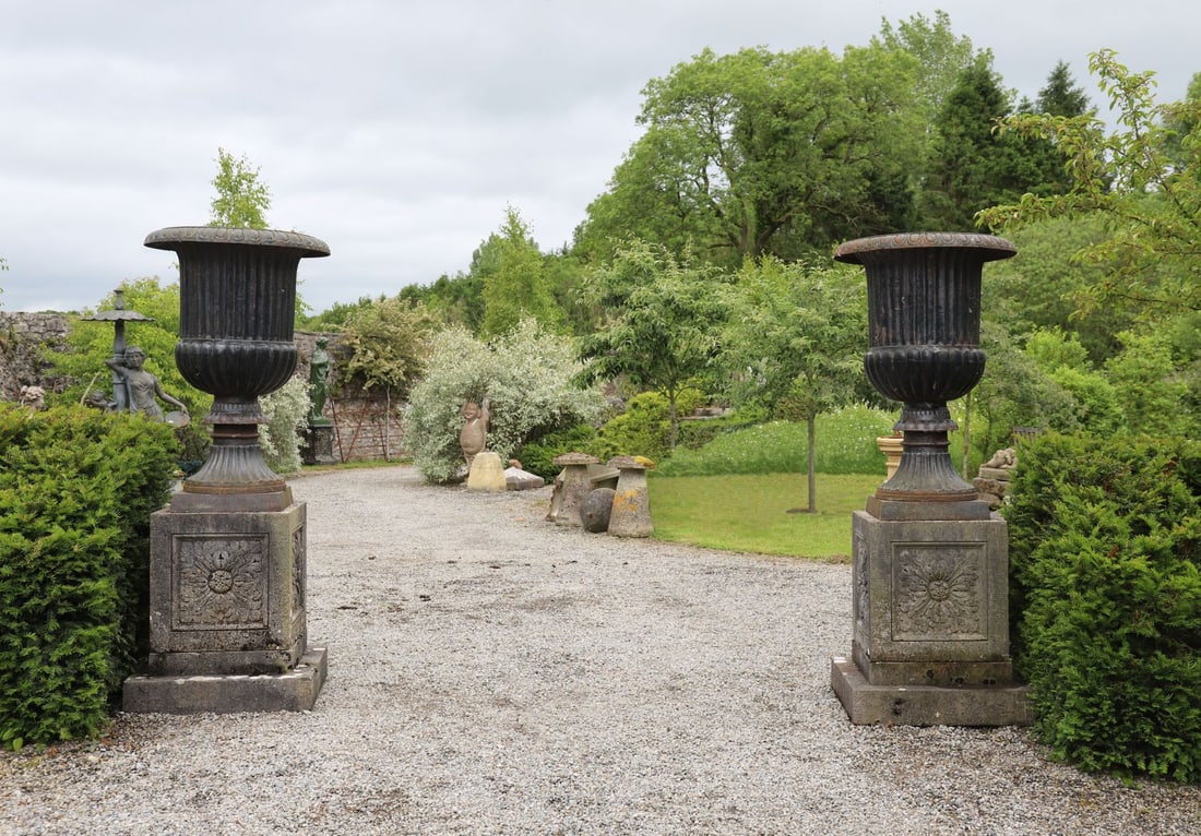 PAIR OF ESTATE CAST IRON CAMPANA URNS: ON LIMESTONE PLINTHS Pair of cast iron campana urns with fluted bodies and gadrooned rims, each raised on a carved limestone pedestal plinth with moulded base. Weathered throughout with attractive gar