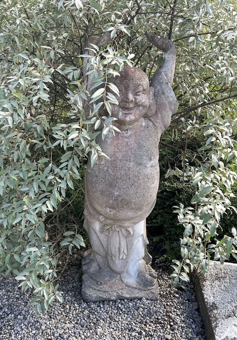 ORIENTALIST COMPOSITE STONE GARDEN FIGURE: Composite stone garden figure depicting a smiling Orientalist male figure standing with arms raised above his head, wearing a draped loincloth and raised on a naturalistic base. Weathered surface pati