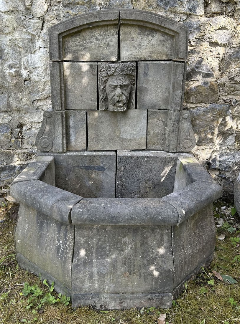 19TH-CENTURY CUT STONE GREEN MAN WALL FOUNTAIN: Architectural cut stone wall fountain of arched form, constructed in sectional blocks and centred by a carved Green Man mask spout beneath a moulded arch. The lower section incorporating a shaped poly