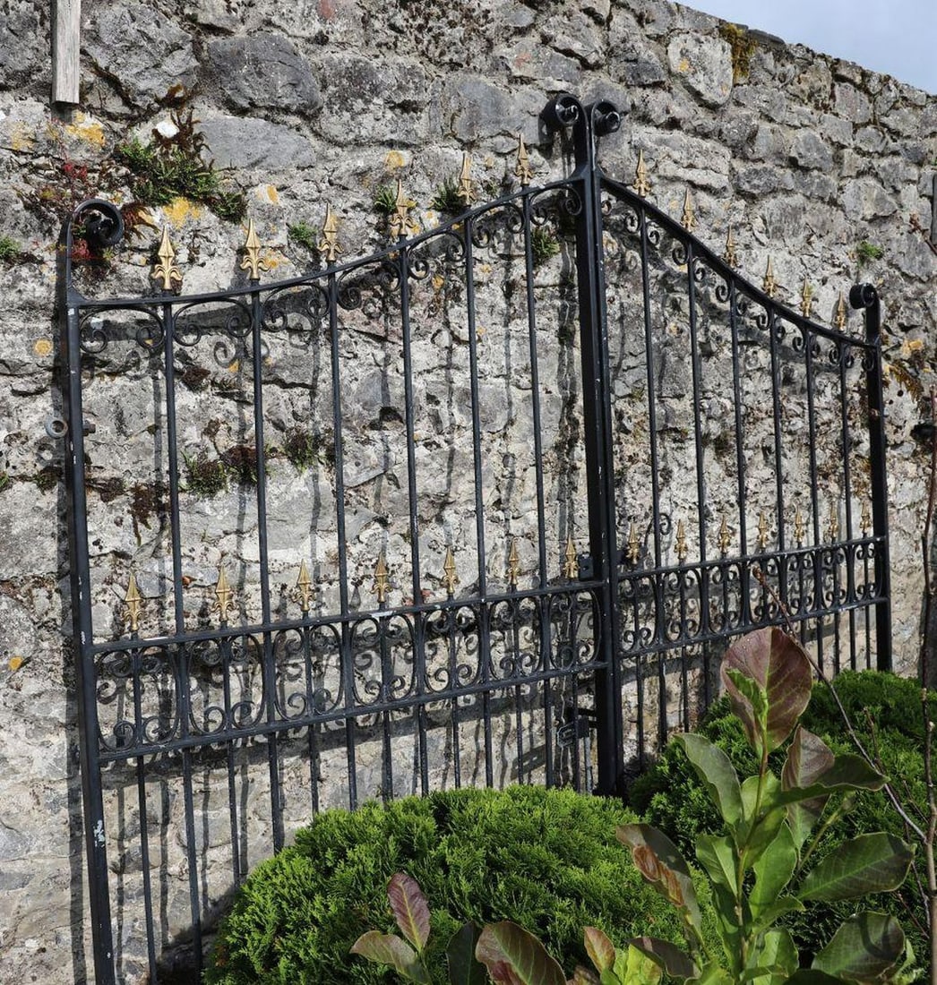 PAIR OF WROUGHT IRON GATES: WITH FLEUR-DE-LIS FINIALS A pair of black painted wrought iron garden or driveway gates, each with scrolled ironwork and vertical bars topped by gilt fleur-de-lis finials. Slightly arched upper frame 
