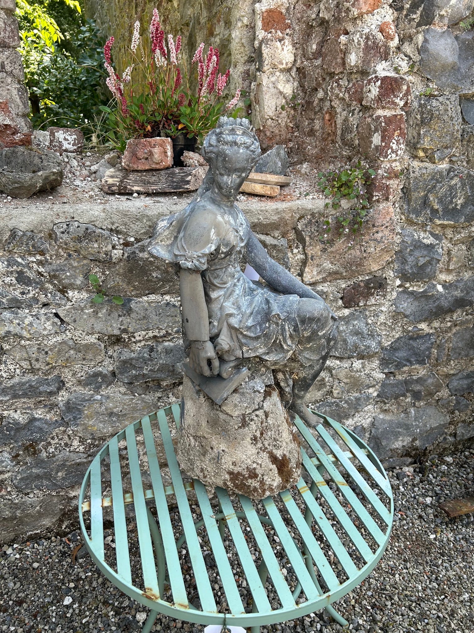 LEAD GARDEN FIGURE OF A CLASSICAL MAIDEN: Lead garden figure depicting a seated classical maiden draped in flowing robes, mounted on a naturalistic stone base with weathered surface patination.