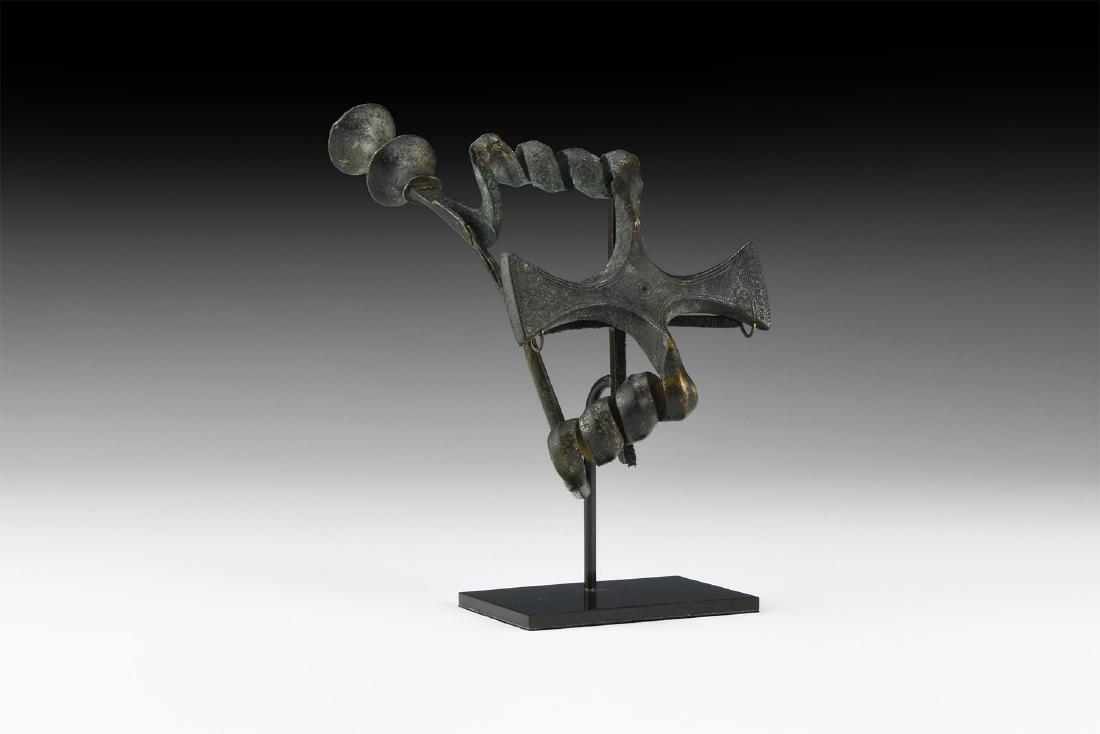 Italic Large Cruciform Fibula: 8th-6th century BC. A large bronze fibula with two coils linking by a cruciform plaque, tapering pin, cottonreel finial with lateral spur forming the catch; incised guilloche and ring-and-dot motifs;
