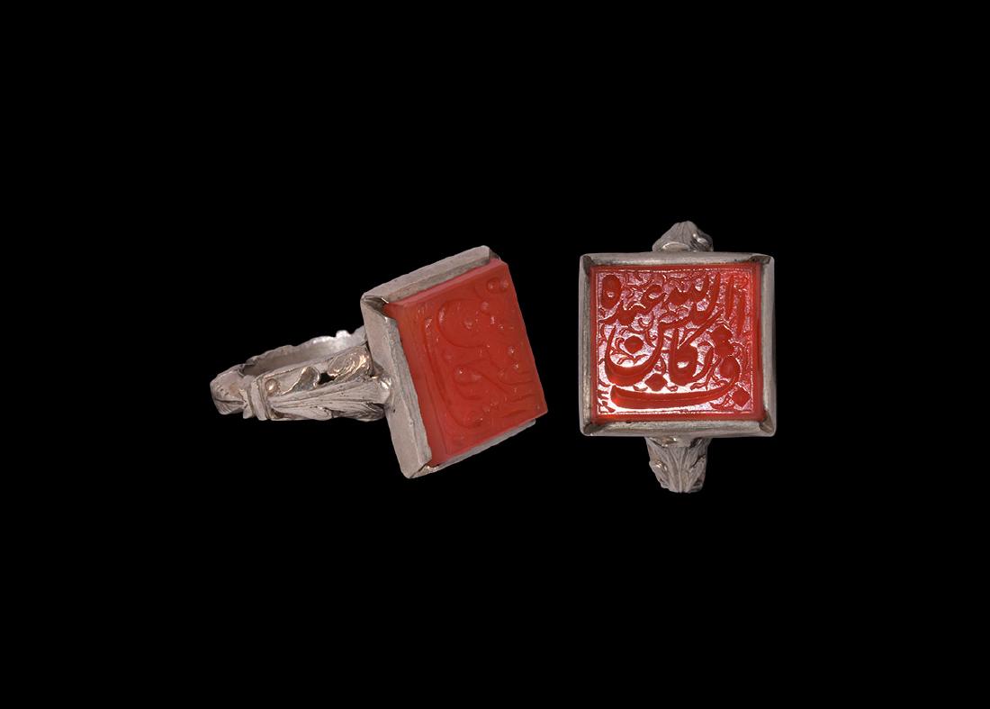 Islamic Calligraphic Jewel in Silver Ring: 19th-20th century AD. A silver ring comprising a hollow-formed D-section hoop with segmented outer face, applied scrolls to the shoulders, square plaque with chamfered edges, inset carnelian cloison w