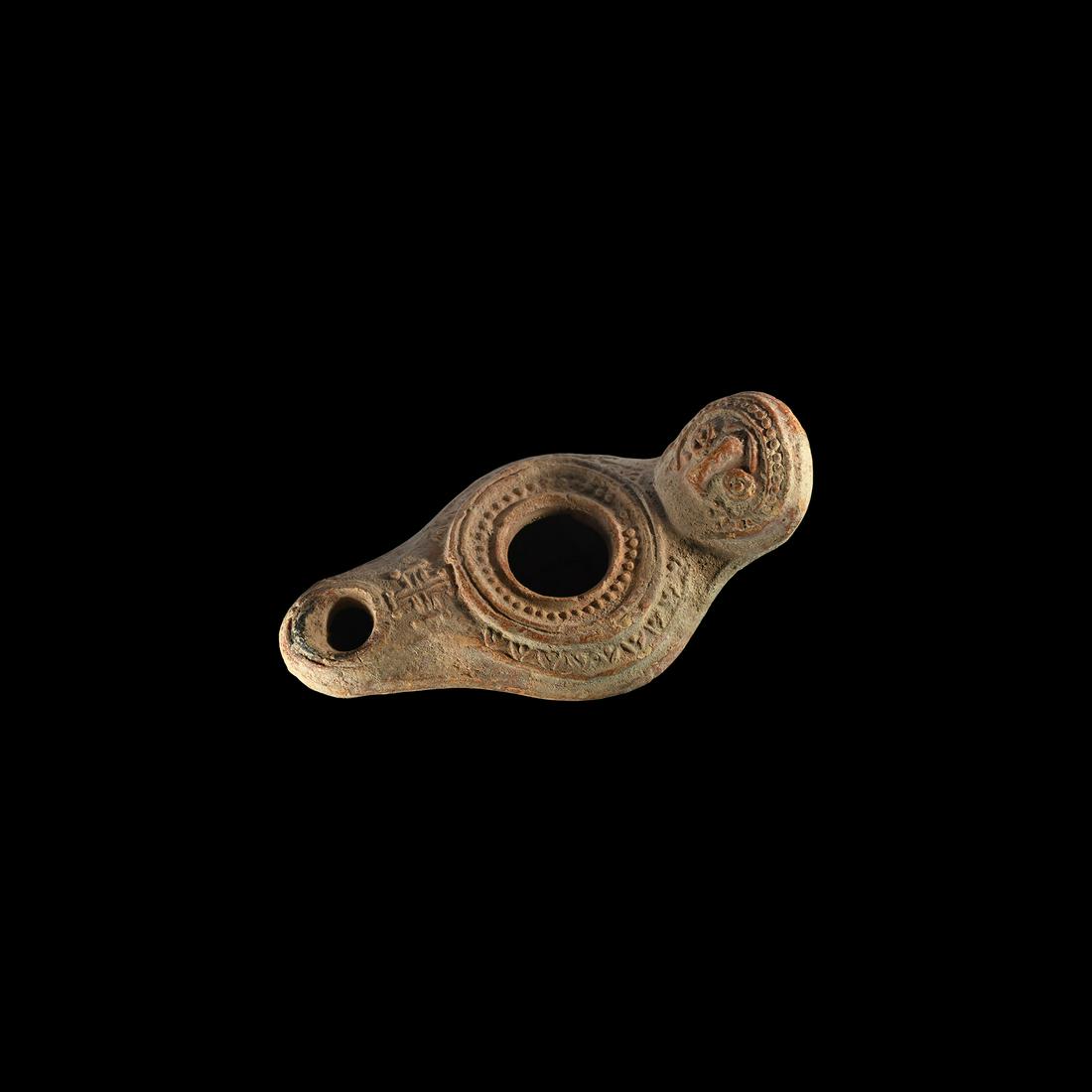 Byzantine Oil Lamp with Face and Cross: 6th-8th century AD. A terracotta oil lamp with tapering nozzle and cross motif, broad discus with pellets surrounding the filler hole, rosette to the underside, reflector with stylised mask and pellet