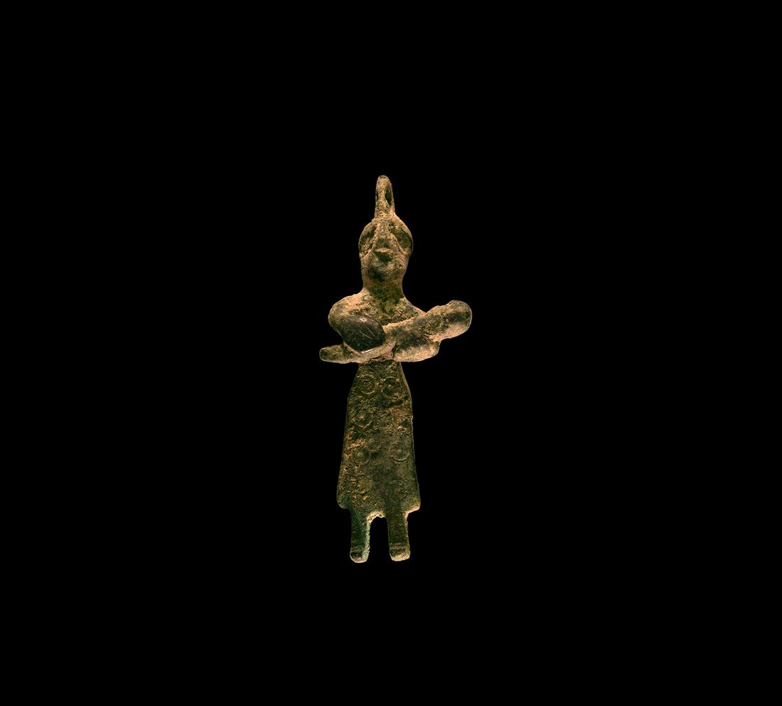 Mother and Child Pendant: 1st millennium BC. A bronze pendant in the form of a standing woman in long robe decorated with eight circle-and-dot motifs, holding a child in both arms, suspension loop to the top of the head. 16 gr