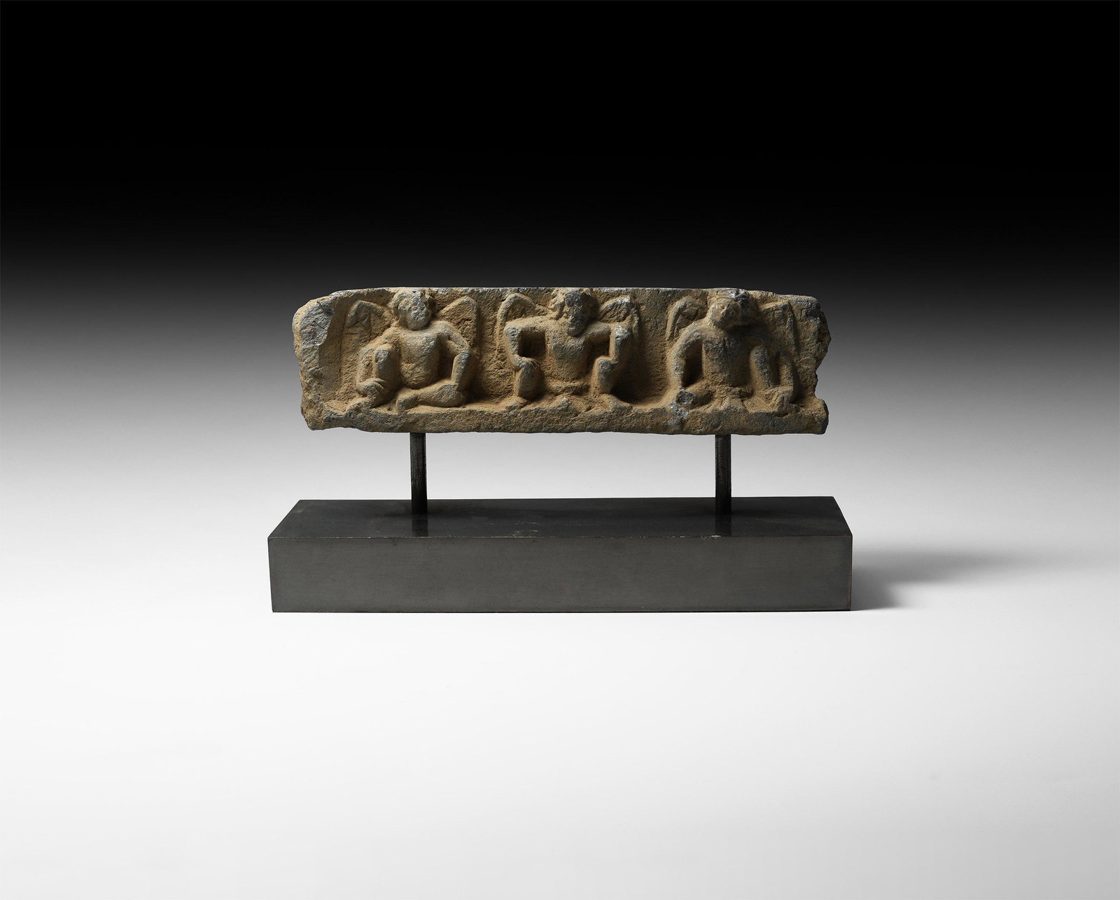 Gandharan Plaque Fragment with Atlantes: 2nd-4th century AD. A carved schist frieze fragment with three crouching atlantes, nude and with wings spread to the rear, on a shallow ledge; mounted on a custom-made stand. Cf. Jongeward, D. Buddhis