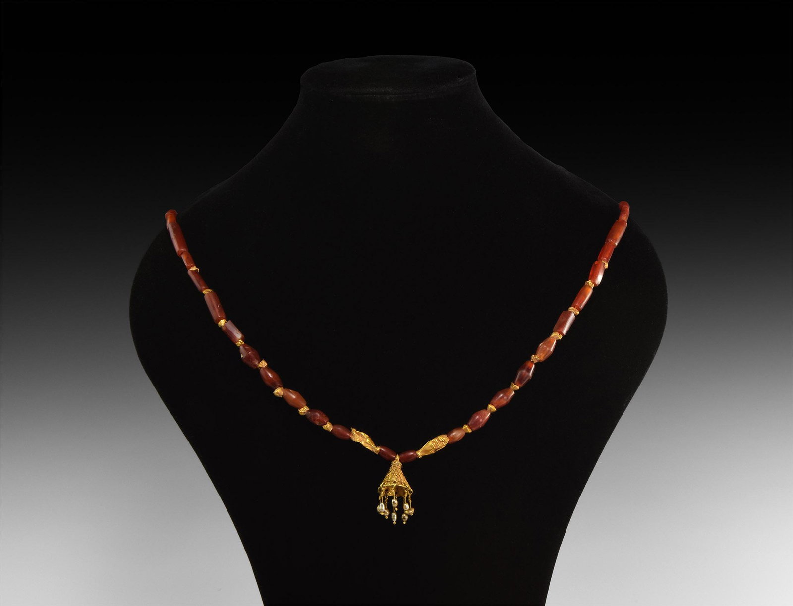 Roman Gold Pendant Necklace: 1st-3rd century AD. A restrung necklace of tubular and biconical carnelian beads with hollow gold spacers, gold bell-shaped pendant with pearl breloques. 30 grams, 45cm (17 3/4"). Property of a Richmo