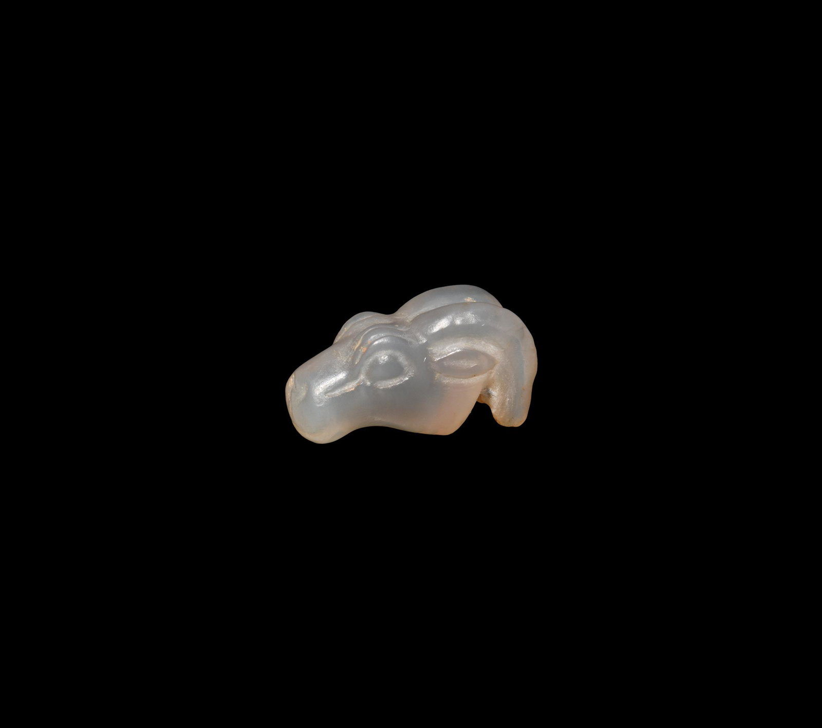 Western Asiatic Ram-Head Amulet: 1st millennium BC. A carved chalcedony pendant of a ram's head with spiral horns and large lentoid eyes; pierced for suspension. 2.4 grams, 18mm (3/4"). Property of a London gentleman; acquired by his