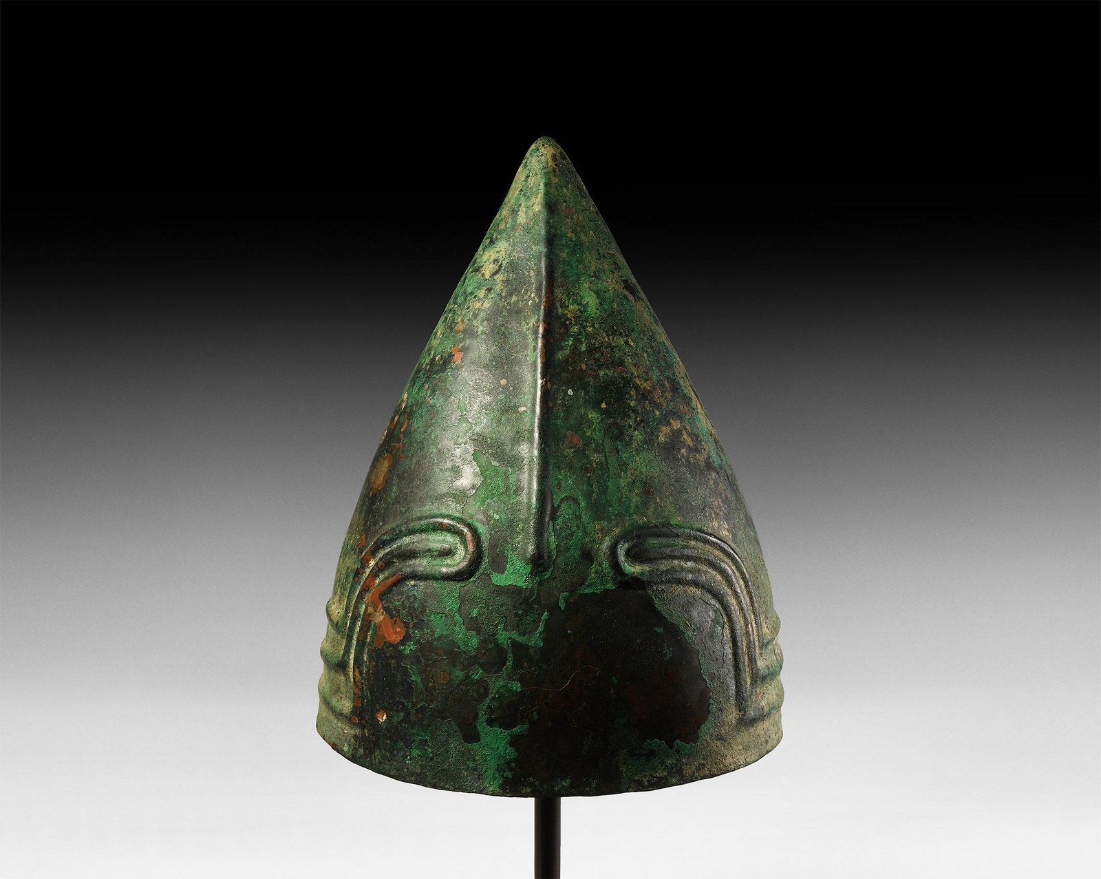 Western Asiatic Decorated Urartian Helmet: 8th century BC. A bronze helmet of conical form made in one piece, a raised groove to the front, three arched ribs with round terminals above the brim resembling eyebrows, chased royal lion motif abov