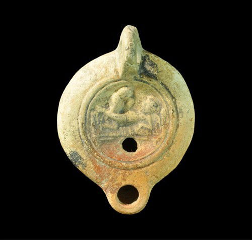 Roman - Erotic Scene Oil Lamp: Circa 1st century AD. A pink and cream oil lamp; on the discus an erotic scene involving a recumbent female and kneeling male within concentric bands; the nozzle plain and handle pierced. Ceramic, 70