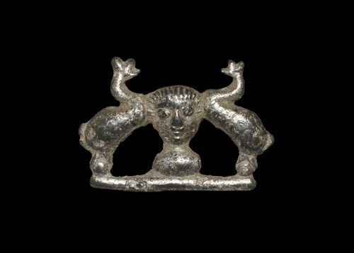 Romano British Cavalry Parade Helmet and Peacocks Plate: Circa 2nd century AD. A silvered bronze plate brooch formed as a pair of opposed peacocks framing a male bust wearing a cavalry parade helmet; pin lost in antiquity. Silvered bronze, 2.36 grams, 23 mm