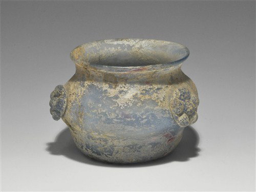 Roman Pale Blue Glass Bowl: Circa 1st century AD. A pale blue glass bowl with carinated profile and everted rim, the base concave; around the circumference three blue glass rosette appliqué motifs; the surface iridescent. Glass