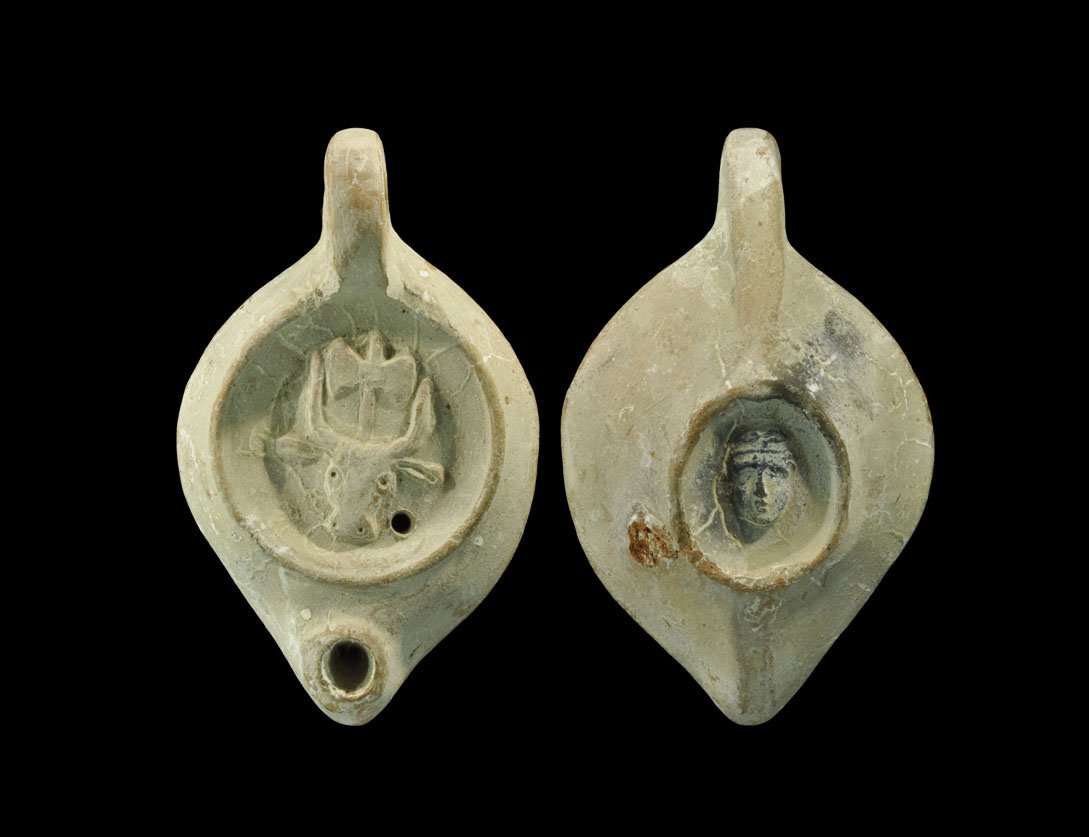 Roman Ceramic 'Theseus and the Minotaur' Oil Lamp: An undated red-brown clay oil lamp with designs on the discus: a bull's head with erect horns and a double-headed labrys axehead between; the handle comma-shaped and pierced; the nozzle plain and coll