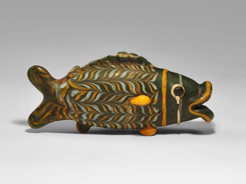 Phoenician Style Core-Formed Polychrome Glass Fish: A undated core-formed glass fish, green-black body with orange and white marvered and applied trails, parts of dorsal fin remaining, standing on feet modelled as fins, hollow. Glass, 49.1 grams, 100 m