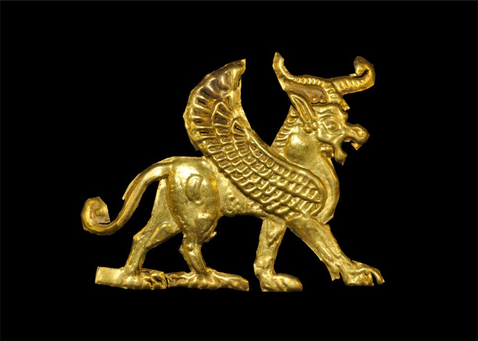 Near Eastern Gold Winged Bull Mount: 1st millennium BC. A repousse gold foil mount in the form of an advancing winged bull with curved horns, gaping mouth, elliptical ear, thick trunk, broad wing and curled tail; to the reverse, a gold a
