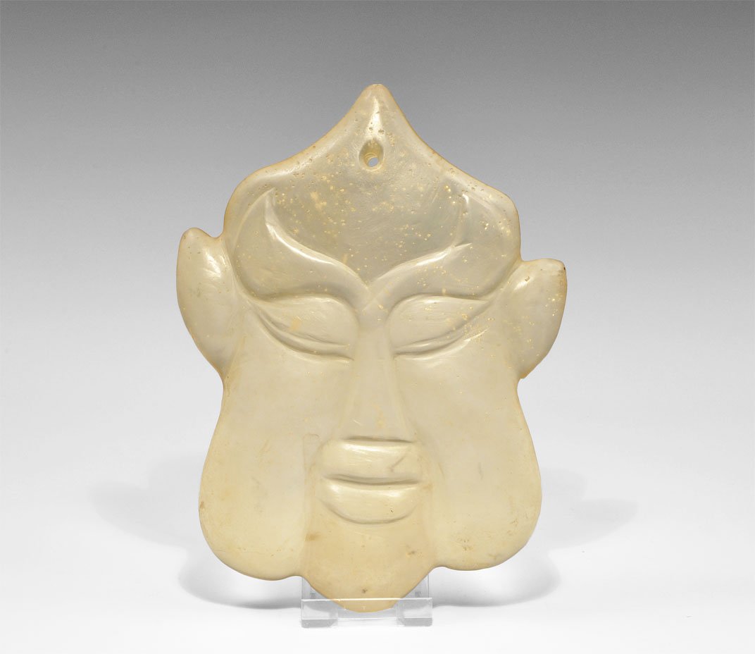 Tibetan Glass Mask Plaque: An undated cast glass plaque depicting a male face with lentoid ears and eyes, thick lips and pointed crown; pierced close to top for attachment. Acquired on the London art market in the 1960's. Glass