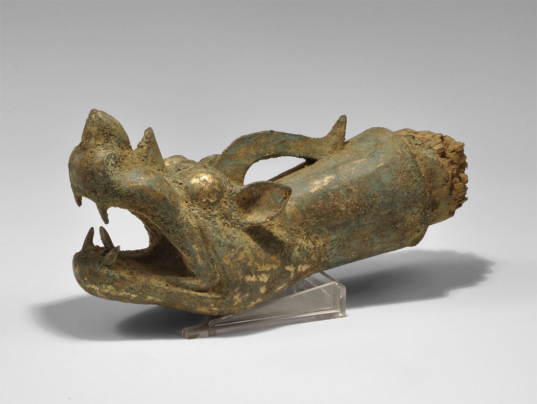 Chinese Gilt Bronze Ordos Style Dragon-Headed Shaft Ter: An undated hollow-cast terminal in the form of a dragon's head with gaping mouth, raised tongue, exposed fangs, bulbous eyes and curled lappet; the conical socket retaining the wooden shaft. From an i