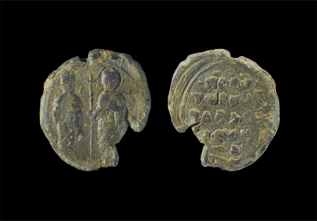 Byzantine Lead Bulla Seal: Circa 12th-14th century AD. A cast lead bulla seal depicting to one face a pair of nimbate figures with a cross between; to the other face, a raised inscription in four lines 'H..O/TWS.A/.DLA./.VE..' 