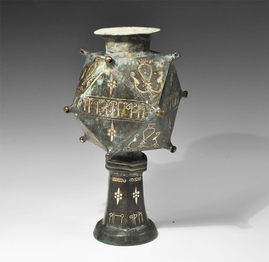 Islamic Bronze Polyhedral Vessel: An undated bronze vase with inlaid surface; the base conical with everted rim, the body formed as a hollow polyhedron of six square and eight triangular sides with arabesque motifs in the triangles an