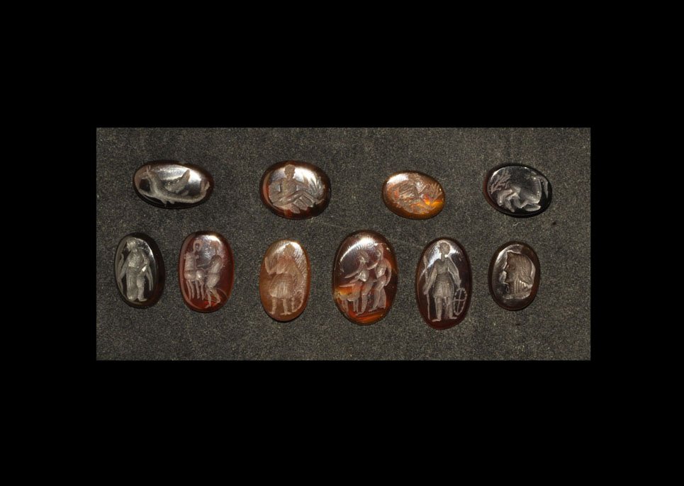 Roman Style Carnelian Intaglio Group: A mixed group of undated intaglios comprising: one winged serpent; one warrior with shield and spear; one fleeing stag with vegetation; one standing robed figure with stalk; two seated figures with ra