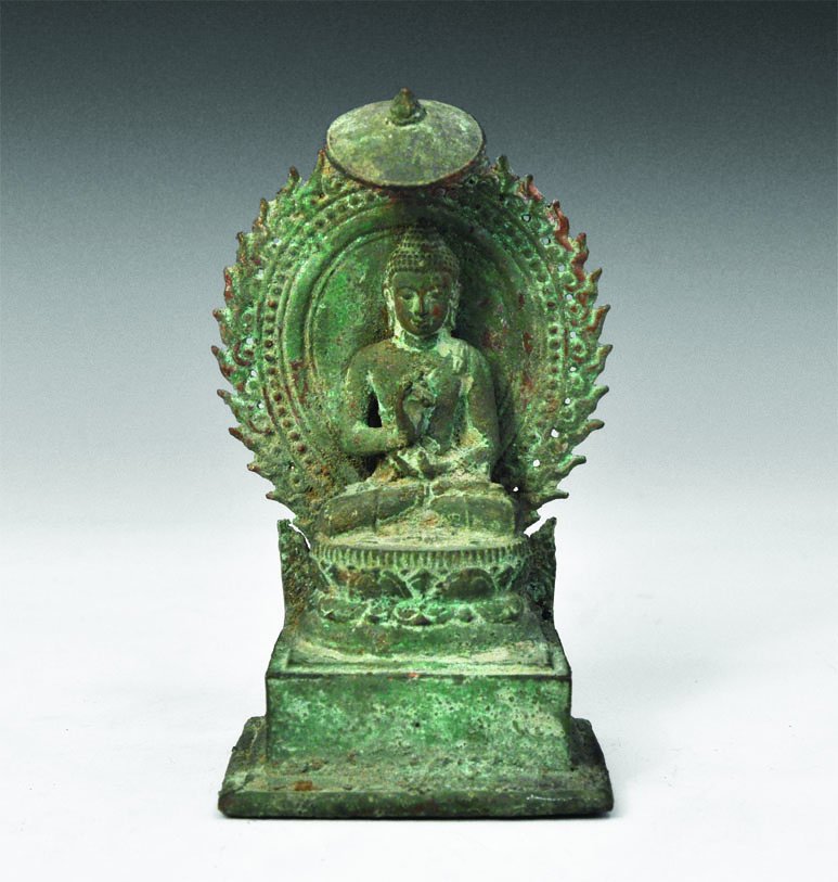Indian Bronze Deity Statuette: An undated cast bronze figure of the Buddha in 10th century style seated on a lotus-leaf column, cross-legged with a pot cradled in the lap in the left hand, the right hand raised in the gesture of pe