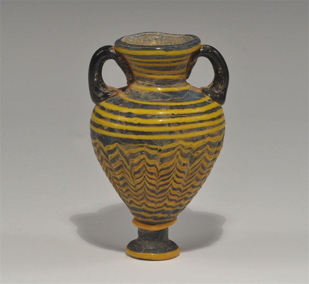 Near Eastern Phoenician Style Miniature Glass Amphora: An undated glass miniature amphora comprising pedestal foot, ribbed piriform body, everted rim and two loop handles, in dark blue with yellow trails. 63 grams, 85 mm (3 1/4"). UK art market, acquired