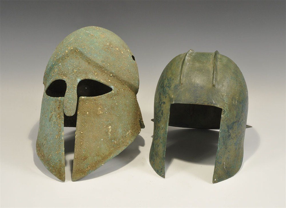 Greek Style Bronze Helmet Group: A mixed group of undated bronze helmets comprising: one Corinthian style with broad cheek-plates, lentoid eye-holes and short nasal, carinated to the lower bowl; one Illyrian style with two ribs to th