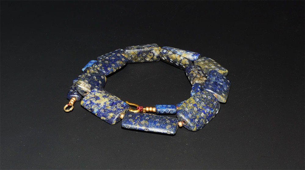 Near Eastern Lapis Lazuli and Gold Bead Necklace: An undated restrung necklace comprising nineteen plano-convex lapis lazuli rectangular beads with incised ring-and-dot detail and interstitial modern gold seed beads; modern silver-gilt clasp. 80 gram