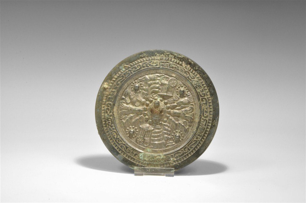 Chinese Bronze Mirror with Horse and Chariot Motifs: Han Dynasty, 206 BC-220 AD A cast disc mirror with one polished surface; to the reverse a central pierced boss surrounded by images of horses pulling chariots, crowned figures and running figures; to 