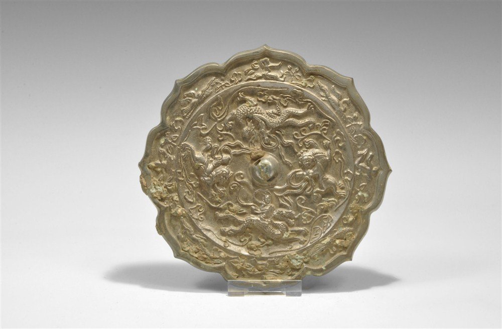 Chinese Bronze Mirror with Rider Motifs: Han Dynasty, 206 BC-220 AD A cast octofoil mirror with one polished surface; to the reverse, a pierced central boss surrounded by four riders on dragons and lions amid scrolled vegetation; to the oute