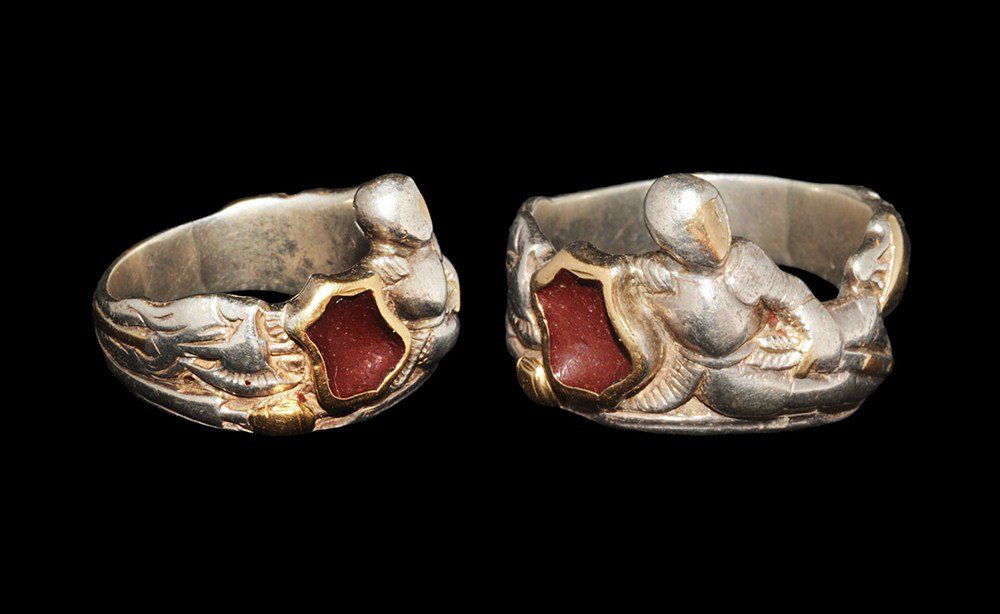 Post Medieval Silver Reclining Knight Finger Ring: Circa 19th century AD. A cast parcel-gilt silver figural finger ring depicting a reclining armoured figure resting on his shield and clutching his sword with his left hand; behind the shield a battle