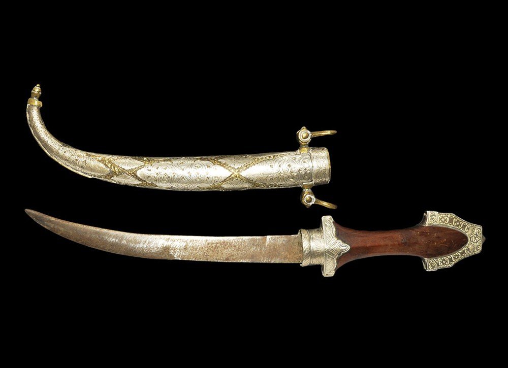 Near Eastern Jambya Dagger and Scabbard: Moroccan, 20th century AD. An ornamental dagger and scabbard; the dagger's curved triangular blade with two ricassos, waisted grip with wooden scales, white metal decorative collar and fittings to the