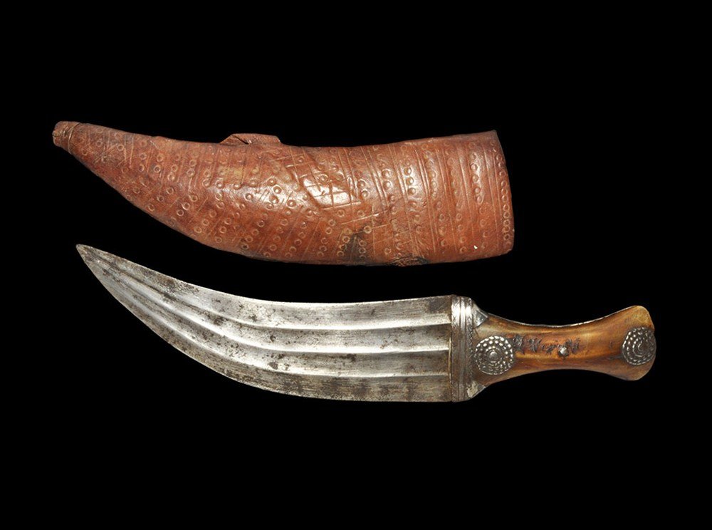 Near Eastern Janbiyya Dagger and Sheath: Omani,19th century AD. A decorative dagger and scabbard; the dagger with three ribs to each face of its curved blade, waisted grip with wooden scales, sheet silver sleeve to one face, four rosette app